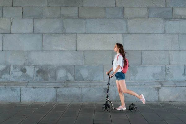 Woman in summer in city on scooter, rides motion. Backpack bag, white shirt pink backpack. Background wall transition by road. Free space for text. Concept of ecological transport healthy lifestyle. — Stock Photo, Image