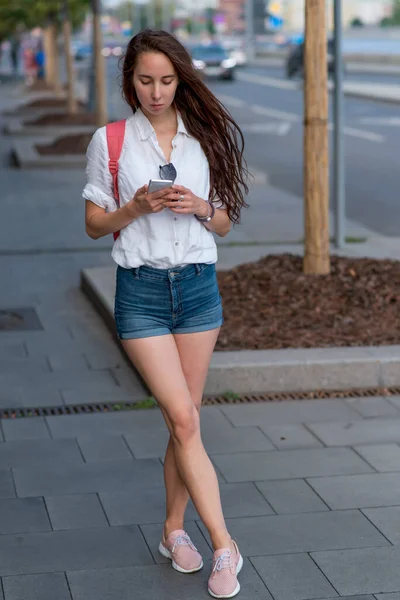 Woman in summer in city in casual clothes shorts shirt backpack, reads and writes a message in chat, online application in a smartphone. Calling a taxi by road, background of roadside by highway. — Stok Foto