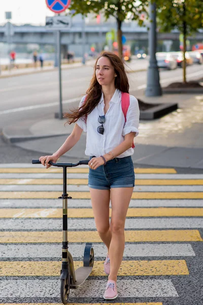 Beautiful woman with lush and long hair, in summer in city crosses road crossing on zebra. Scooter shorts and shirt with pink backpack. Traffic safety concept. – stockfoto