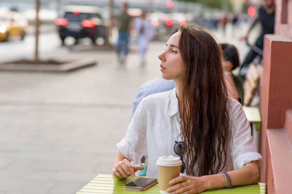 Beautiful woman sits in street cafe, listens voice message online application on Internet, city, waiting friends, meeting date. Free space for a copy of text. Cup with coffee, tea, breakfast, lunch. — Stok Foto