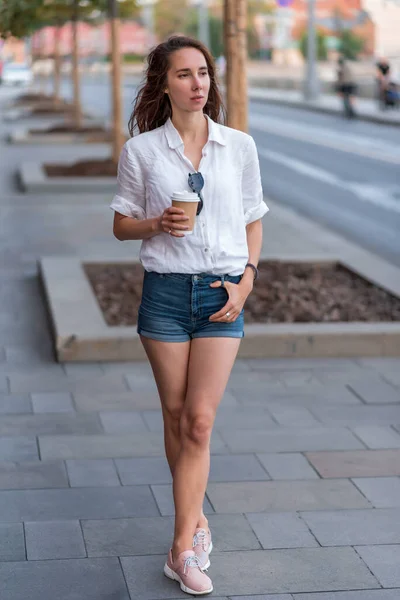 Beautiful woman in summer in city holds a cup of coffee and tea in her hand, waiting for friends, meeting and date. Breakfast lunch and break. Casual wear denim shorts, white shirt. — Stok Foto
