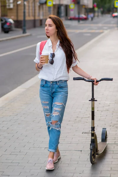 Beautiful woman holds a glass of coffee and tea in her hand, walks with a scooter. Breakfast lunch and break. Casual wear jeans, white shirt. It goes in summer, along road. — Stok fotoğraf