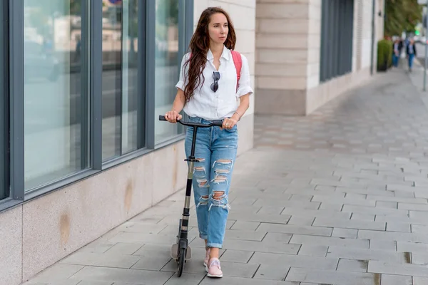 Beautiful woman with long hair walks in city in summer, holds a scooter in her hands, background is road sidewalk glass windows of showcase of building. Free space for a copy of text. — Stok Foto