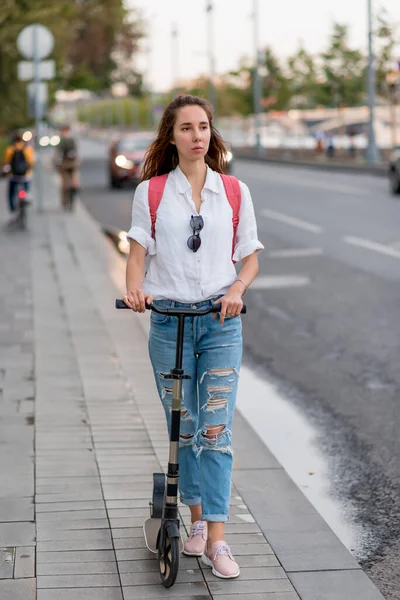 Beautiful woman with long hair walks in city in summer, holding a scooter in her hands, background is road sidewalk cars and cyclists. It goes in summer, along the road. — Fotografia de Stock