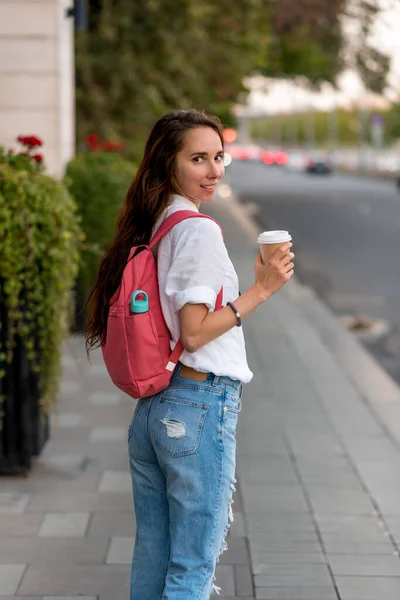 Красивая девушка, счастливая, улыбается, радуется новой встрече с незнакомцем, держит в руках чашку кофе, разворачивается летом в городе. джинсы в белой рубашке, розовый рюкзак. брюнетка. — стоковое фото