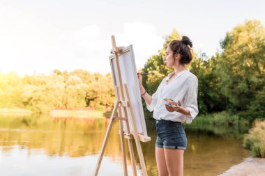 Kız sanatçı, kadın manzara resmi çizer, yaz gölü göleti göleti, beyaz gömlek kot şortu, yaratıcılık sanatını yaratır. Boş beyaz tuval. Arka plandaki ağaçlar su. Fırça rengi paleti boyaları.