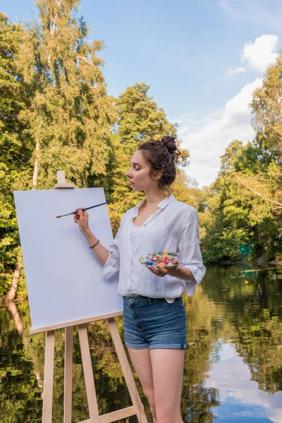 Wanita menggambar sebuah gambar, gadis seniman, taman musim panas danau sungai, kemeja putih denim celana pendek, menciptakan kreativitas dan suasana artistik. Kanvas putih kosong, memulai. Latar belakang pohon air. — Stok Foto