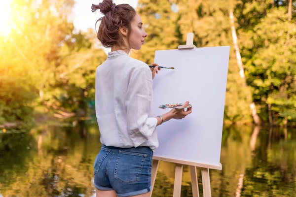 Wanita menggambar gambar, melihat ke belakang, gadis seniman, taman musim panas danau kolam sungai kemeja putih denim pendek, menciptakan kreativitas artistik suasana hati. Kanvas putih kosong, memulai. Latar belakang pohon air. — Stok Foto