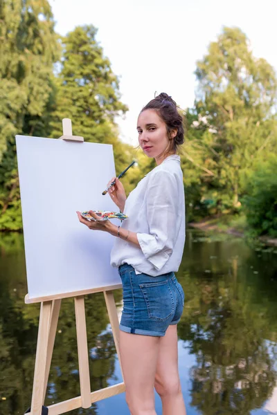 Wanita menggambar gambar, gadis artis, musim panas di taman danau dan kolam sungai, kemeja putih denim celana pendek, menciptakan kreativitas artistik suasana hati. Latar belakang pohon air. Palet kuas dan warna dengan cat. — Stok Foto
