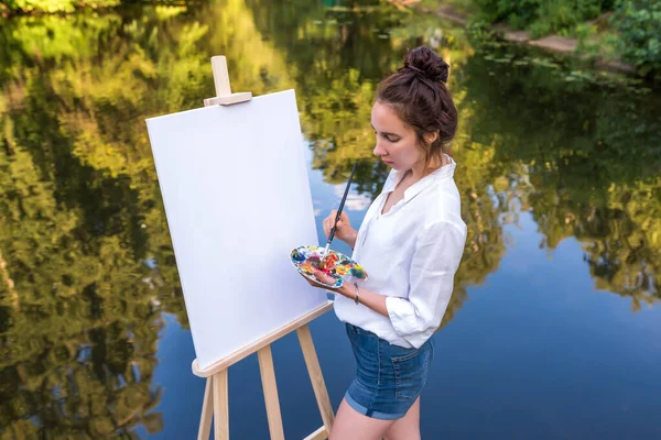 Wanita menggambar gambar, gadis seniman, taman musim panas danau kolam sungai, kemeja denim, menciptakan kreativitas artistik suasana hati. Kanvas putih kosong, memulai. Latar belakang air. Cat palet warna kuas. — Stok Foto