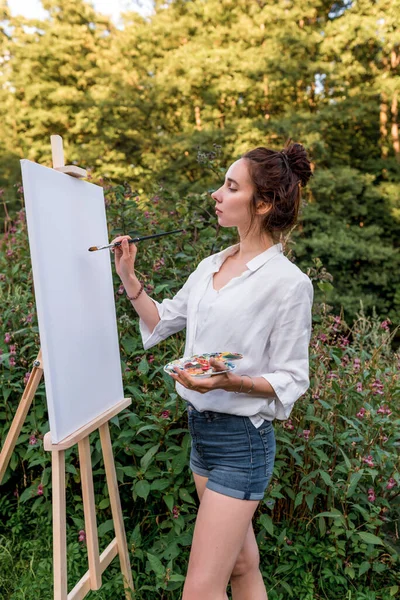 woman paints picture landscape, girl artist, summer forest, creating creativity artistic mood. Blank white canvas, getting started. Grass trees background. Brush color palette with paints.