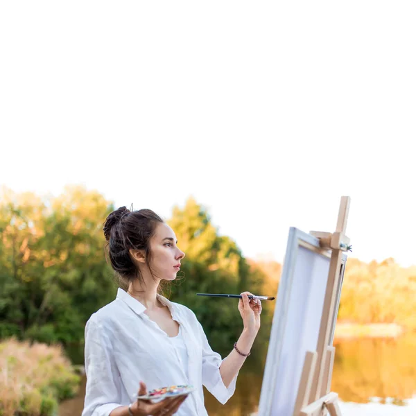 White background free for text, woman draws picture of landscape, in summer pond of lake, white shirt, creating a creative artistic mood. Brush and color palette with paints. — Stock Photo, Image