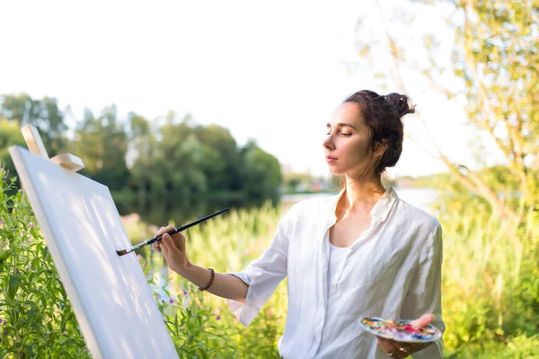 Close-up, woman paints picture landscape, summer pond lake, girl artist, white shirt, creating creativity artistic mood. Blank white canvas, getting started. Background trees with water.