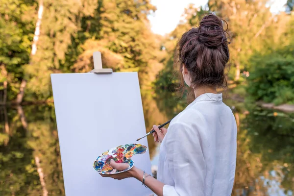 Frau malt Bild Landschaft, Mädchen Künstler, Blick zurück, Sommer Park Teich Fluss, weißes Hemd, die Schaffung von Kreativität künstlerische Stimmung. Leere weiße Leinwand, los geht 's. Pinsel Farbpalette Farben. Stockbild