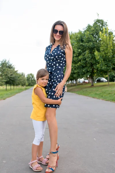 Dítě dívka objetí žena maminka, procházky s malou holčičkou dcera 5-7 let, v létě v městském parku, ležérní oblečení a šaty, šťastná procházka, úsměv a relaxovat o víkendu. — Stock fotografie