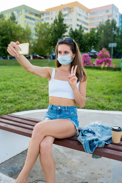 Θηλυκό γυναίκα στην πόλη το καλοκαίρι στην ιατρική μάσκα, κάθεται σε πάγκο εικόνες smartphone, online εγγραφή στο Διαδίκτυο, τηλέφωνο εφαρμογή, κλήση βίντεο λευκό θέμα casual ρούχα τζιν σακάκι, φλιτζάνι τσάι καφέ. — Φωτογραφία Αρχείου