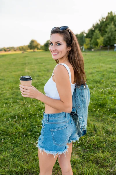 Gelukkige vrouw in de zomer in het park houdt beker met koffie thee handen, weiden, geniet van rust. Emoties van tederheid, plezier, ontbijt in de natuur. Witte tank top gelooid leer, denim shorts en jas. — Stockfoto