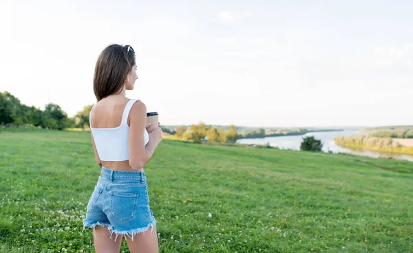 Gadis atletis yang cantik di luar ruangan musim panas, memegang cangkir dengan teh kopi di tangan, kesenangan relaksasi, salinan ruang kosong teks. Celana pendek denim tank putih. Sisi tampilan. Danau sungai rumput latar belakang. — Stok Foto