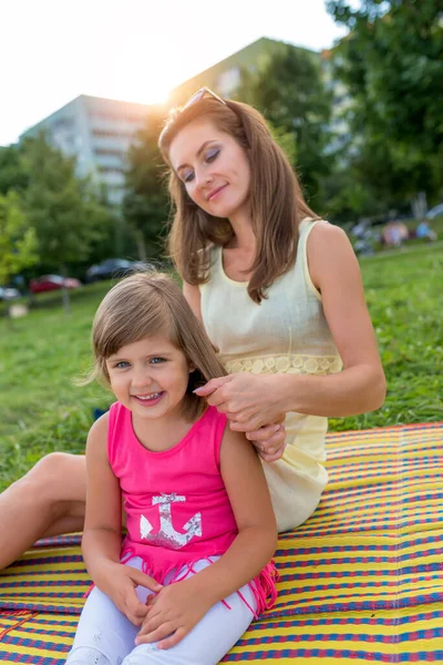 Frau Mutter im Sommer im Park sitzt auf einer Decke, richtet ihr Haar für ein kleines Mädchen Tochter 5-6 Jahre alt, Emotionen der Freude, Spaß und Entspannung, Pflege der Haare, glückliches Familienkonzept. Stockbild