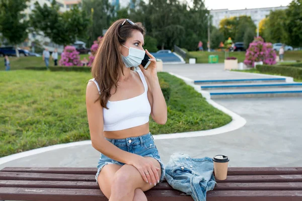 Mujer mujer ciudad verano máscara médica, se sienta en el banco, taza de té de café. Habla por teléfono, escucha mensaje de voz, Internet en línea, aplicación en el teléfono, chaqueta de mezclilla de ropa casual de tema blanco, Fotos de stock libres de derechos