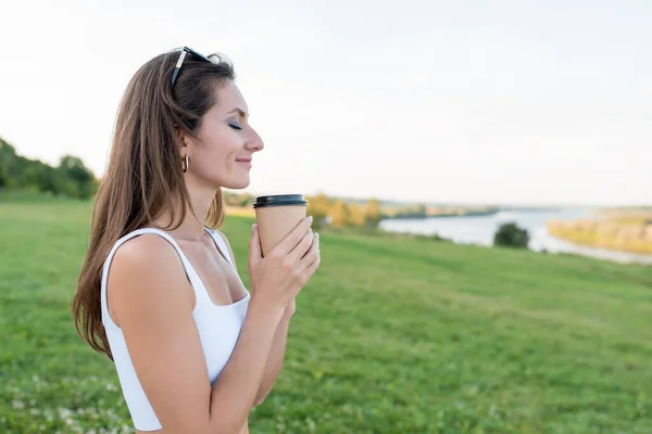 夏の公園で幸せな女性が手にカップコーヒーティーを保持し、テキストコピーのための無料のスペース、風景の牧草地、残りのにおいを楽しむコーヒー茶を飲む。優しさ、自然の中での喜びの朝食の感情. ロイヤリティフリーのストック画像