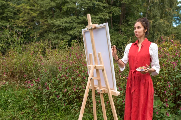Wanita cantik dengan baju merah kemeja putih, hutan taman musim panas, menggambar, menciptakan lanskap teks salinan ruang kosong. Kreativitas dan konsep seni. Latar belakang pohon dan semak-semak. — Stok Foto