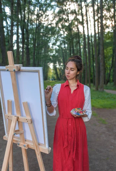 Wanita cantik bergaun merah di musim panas di taman dan hutan, menggambar, menciptakan kreativitas dan kreativitas pemandangan. Kreativitas perempuan dan konsep seni. Latar belakang pohon dan semak-semak. — Stok Foto