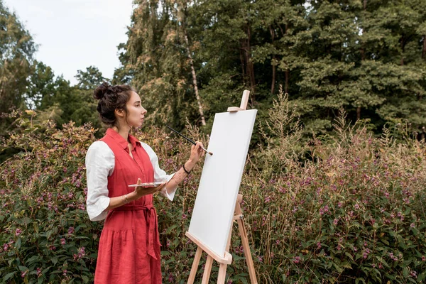 Piękna kobieta w czerwonej sukience biała koszula, w letnim lesie parkowym, rysuje obraz, białą płócienną farbę i pędzel, wolne miejsce na kopię tekstu. Koncepcja kreatywności artystycznej. Obrazek Stockowy