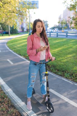 Yazın şehirde scooter süren bir kadın. Ayakta duruyor ve telefonla güzergaha bakıyor. Yol güvenliği kurallarına uymak. Pembe ceket ve kot pantolon. Arama ve internet navigasyon uygulamasını yönlendir.