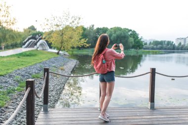 Yaz mevsiminde bir kadın gölün, gölün, gölün, gölün yakınındaki bir su deposunda akıllı telefondaki video fotoğraflarını kaydediyor. Gezmek ve seyahat etmek. Arkadan bak. Sırt çantalı Denim şort ceket.