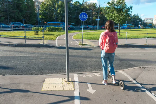 Fata pe scuter vara în oraș. Stă cu spatele, la răscruce de drumuri, aşteptând să treacă maşina. Copie gratuită a textului. Respectarea normelor de siguranță rutieră. Geanta roz si blugi. Biciclete traseu semn . — Fotografie, imagine de stoc