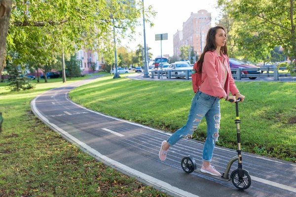 Femeie pe un scuter, vara în oraș plimbări în mișcare, spațiu liber pentru o copie a textului. Jacheta roz, rucsac, blugi. Fundal gazon verde, copaci, traseu de biciclete, drumuri și mașini . — Fotografie, imagine de stoc