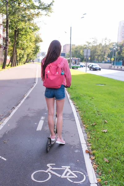 Donna guida uno scooter in città in estate, una vista da dietro, pantaloncini di jeans e una giacca rosa con uno zaino. La pista ciclabile è bagnata dopo la pioggia. Auto fondo strada. — Foto Stock