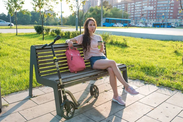 Nyáron egy nő a városi parkban, munka után pihen, kezében egy csésze kávé teával. Pihenés és ebéd munka után, reggeli szünet. Zöld fű háttér és autók, robogó. — Stock Fotó