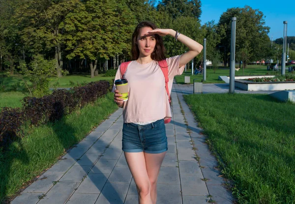 Femme en été dans un parc de la ville regarde au loin, se couvre avec sa main du soleil, une réunion et une date, une tasse de thé au café dans sa main. Belle fille en vacances. — Photo