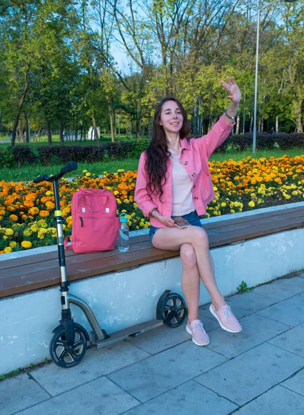 Wanita di musim panas di taman kota melambaikan tangannya, tersenyum gembira, bertemu dan kencan, scooter shop tas dan botol dengan air. Gadis cantik berlibur.. — Stok Foto