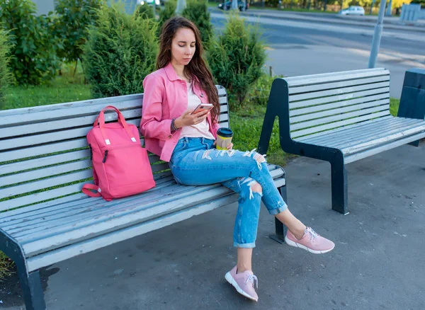 Frau in der Stadt auf Bank im Sommer, liest schreibt Nachricht im Chat, soziale Netzwerke im Internet, Anwendung Smartphone. Casual wear, rosa Jacke, Bag Jeans. Entspannung von Kaffee-Tee nach der Arbeit. Stockfoto