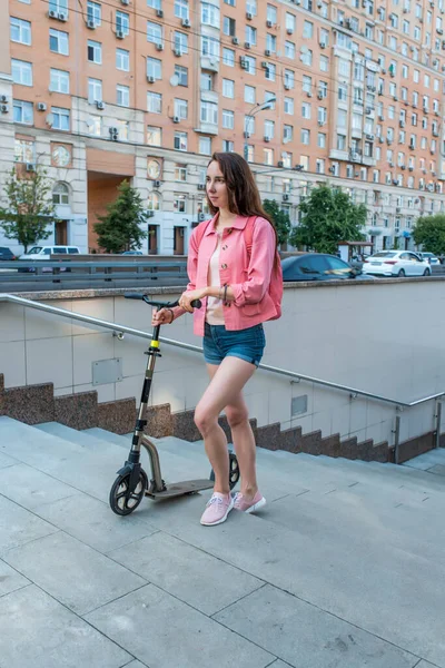 Mulher no verão na cidade vai à passagem subterrânea com uma scooter, calças curtas de ganga e uma jaqueta rosa com uma mochila. Cumprimento das regras de segurança rodoviária. Carros rodoviários de fundo e edifícios. Fotos De Bancos De Imagens