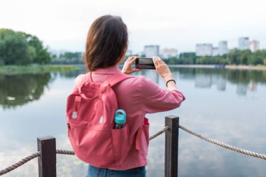 Yakın plan, arkadan manzara, nehir gölü yaz göletinde bir kız, akıllı telefondaki video ve fotoğrafları kaydediyor. Seyahat gezisi. Manzara fotoğrafçılığı. Kot şortlu pembe ceketli, sırt çantalı..