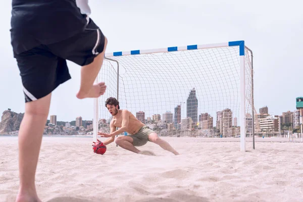 Two boys play soccer on the beach in summer, one shoots the ball in out-of-focus foreground and the other stops the shot before the ball enters the goal.