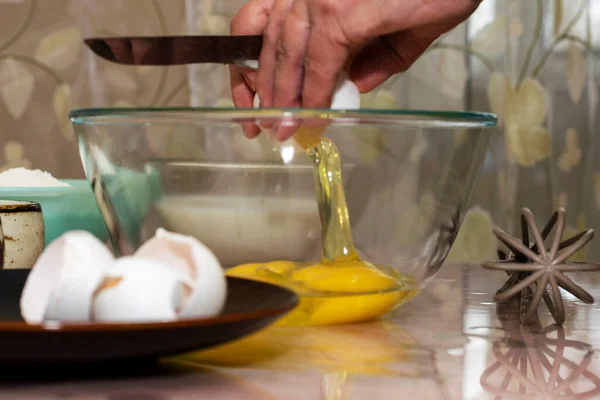 An elderly woman breaks an egg with a knife into a glass bowl for making pancake batter.Ingredients on the table-milk, salt, sugar, wheat flour, butter.Selective focus.