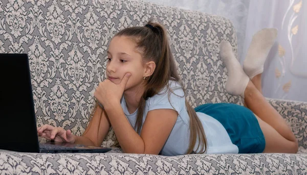 Egy gyönyörű tinédzser lány fekszik egy szürke ágyon egy laptoppal, és online órákat vesz.. — Stock Fotó
