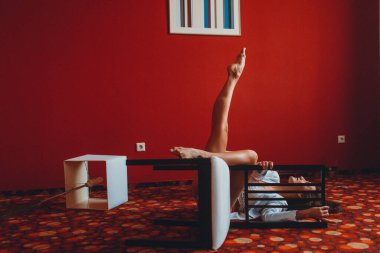 Beyaz ceketli ve kırmızı mayo giyen genç bir kız, kırmızı bir odada esneme ve denge ile meşgul mobilya ve zemin lambaları ile