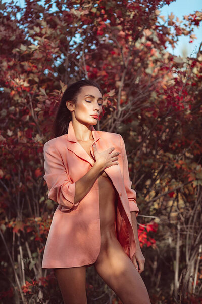 Young beautiful brunette girl with good makeup on her face with expressive features and figure. walks in the fall among beautiful red trees, she is wearing a stylish pink suit and she is a little naked