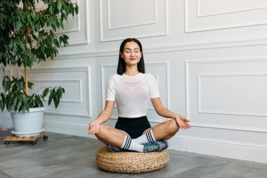 Asyalı görünümlü güzel bir kız, evde spor yapıyor, açık renk bir üst ve siyah bir kıç giyiyor. Çok olumlu düşünüyor.