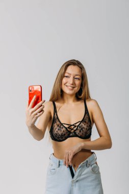 Genç ve güzel bir kız. Saçları siyah iç çamaşırlı ve kot pantolonlu. Stüdyoda beyaz arka planda bir telefonla selfie çekiyor.