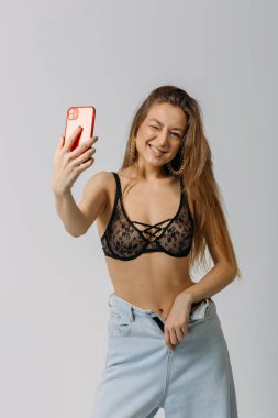 Genç ve güzel bir kız. Saçları siyah iç çamaşırlı ve kot pantolonlu. Stüdyoda beyaz arka planda bir telefonla selfie çekiyor.