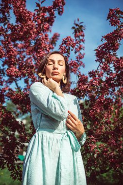 Genç ve güzel bir kız parkta çiçek açan bir elma ağacının altında duruyor. Çok güzel bir açık renk elbise giyiyor..