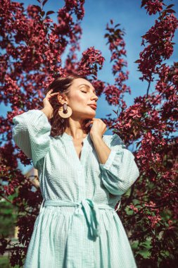 Genç ve güzel bir kız parkta çiçek açan bir elma ağacının altında duruyor. Çok güzel bir açık renk elbise giyiyor..
