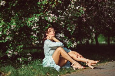 Genç ve güzel bir kız parkta çimlerin üzerinde çiçek açan beyaz bir elma ağacının altında oturuyor, açık renk bir elbise giyiyor..